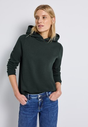 Dunkelgrüner Hoodie aus weichem Stoff, mit einer Kapuze mit Kordelzug und gerippten Bündchen. Kombiniert mit blauen Jeans mit doppeltem Knopfverschluss.