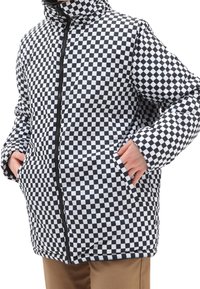 Person iført en sort og hvid ternet pufferjakke med hænderne i lommerne, parret med lysebrune bukser.