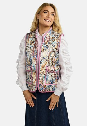 Nasmejana ženska s svetlimi lasmi, oblečena v belo bluzo z nabranimi rokavi in barvno prešito telovnico s cvetličnim vzorcem ter roza obrobo.