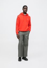 Sudadera con capucha roja con bolsillo frontal, combinada con pantalones grises de corte amplio. Zapatos negros completan el conjunto. Diseño sencillo, con marca mínima.