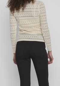 Ljus beige spets långärmad topp med blommönster, bärs över svarta byxor. Toppen har en figursydd design med intrikata texturdetaljer.