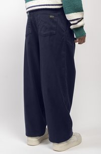 Pantalons larges bleu marine en tissu mélangé de coton, dotés d'une poche arrière avec une étiquette, associés à des baskets blanches.