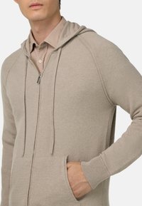 Béžová mikina na zip vyrobená z měkké tkaniny, s kapucí na stažení, žebrovanými manžetami a předními kapsami. Hladká textura s minimálním zdobením.