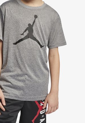 Ung person iført en grå T-shirt med en sort silhuet af en basketballspiller og sorte mesh-shorts med rød grafik og hvid tekst.