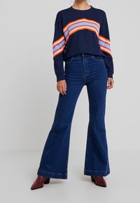 Sudadera navy con un diseño de rayas naranja y azul claro en las mangas, combinada con unos vaqueros azules acampanados y botines de tacón borgoña.