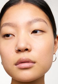 Gros plan sur un visage avec une peau lisse, des yeux marron clair et un anneau en argent au nez. Les traits incluent un maquillage minimal et de petites boucles d'oreilles.