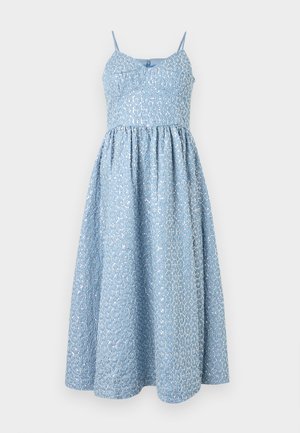 Γυναίκα στέκεται φορώντας ένα μίντι αμάνικο ανοιχτό μπλε φόρεμα με λεπτές τιράντες και μαύρα ανοιχτά πέδιλα με χαμηλό τακούνι σε απλό φόντο.