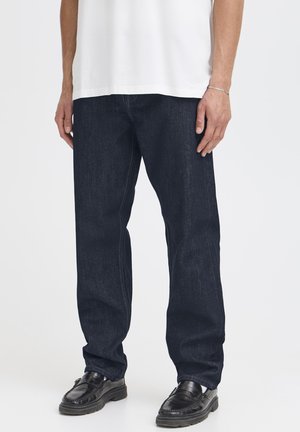 Mand iført mørkeblå jeans med lige ben, sorte læder loafers, hvide sokker, en hvid skjorte og et sølv armbånd på højre håndled.