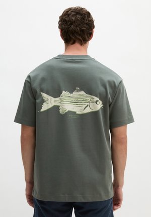 Mann mit lockigem Haar, der ein grünes T-Shirt mit einem großen Fischmotiv auf dem Rücken trägt.