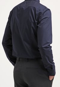 Camicia a maniche lunghe blu navy con una texture liscia, colletto classico e polsini con bottoni, abbinata a pantaloni scuri. Dettagli di design sottili visibili.