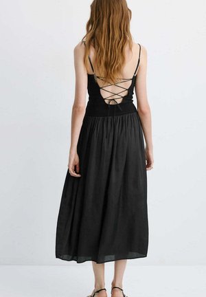 Femme aux longs cheveux portant une robe noire sans manches avec un dos lacé croisé ouvert, debout devant un fond clair uni.
