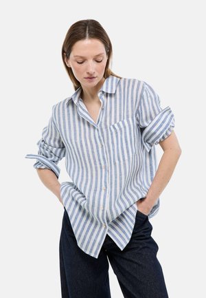 Femme portant une chemise à rayures verticales bleues et blanches avec les manches retroussées et un pantalon foncé, debout les mains dans les poches, regardant vers le bas.
