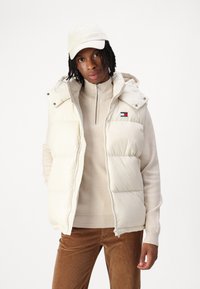 Gilet rembourré en crème clair avec une capuche amovible, fermeture éclair sur le devant et détail de logo. Porté par-dessus un pull beige et associé à un pantalon en velours côtelé marron.