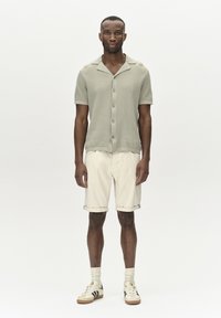 Ljusgrön kortärmad stickad skjorta med krage, tillsammans med krämfärgade shorts. Modellen har på sig vita sneakers med svarta och gyllene detaljer.