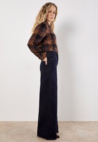 Woman with long wavy blonde hair wearing a brown and black checkered shirt tucked into high-waisted dark blue wide-leg jeans, standing sideways.