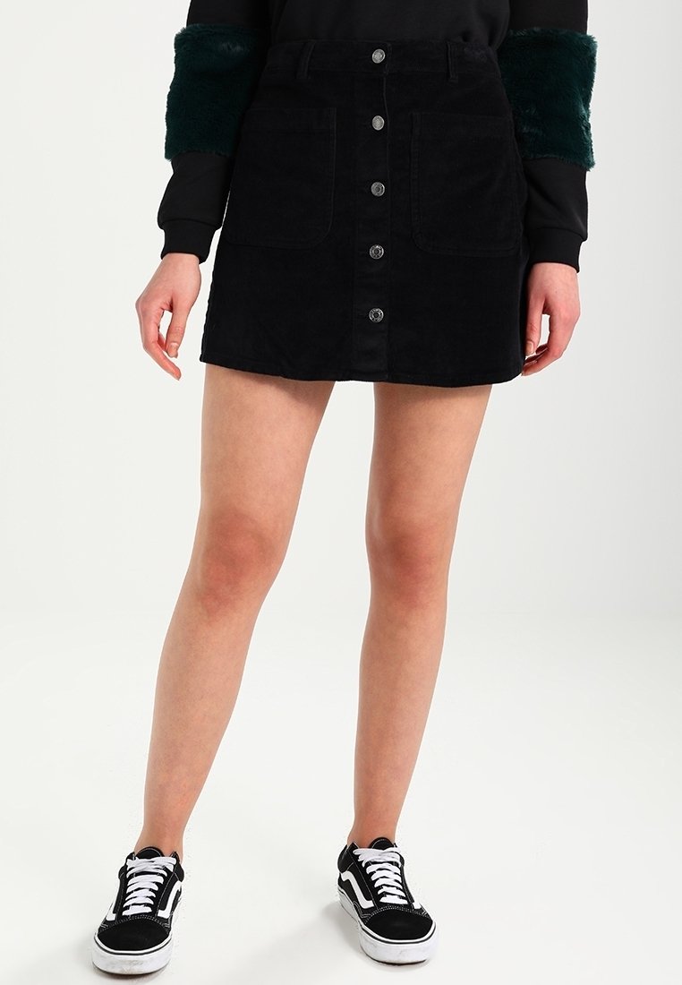 Personne portant une jupe noire boutonnée sur le devant, un haut à manches longues noir et vert, et des baskets noires et blanches, debout devant un fond uni.