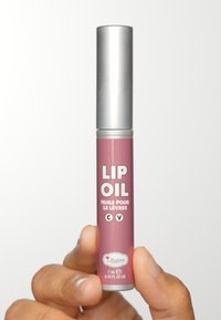 Lip oil i en cylindrisk, matt mauve behållare med ett silverlock. Etiketten har vit text och branding. Storlek: 7 mL. Hålls av en hand.