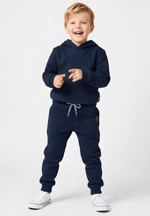 Granatowa bluza z kapturem i dopasowane joggery z dużymi kieszeniami i sznurkiem do regulacji. Bielone trampki z czerwonymi akcentami dopełniają strój.