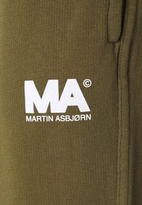 Zielony, oliwkowy materiał dzianinowy z białym napisem "MA MARTIN ASBJØRN" nadrukowanym w pobliżu szwu oraz otwarcia kieszeni.