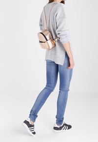 Pieni ruusukultainen reppu, jossa on tyylikäs muotoilu, varustettuna vetoketjulla ja säädettävillä olkahihnoilla, jota käytetään harmaan neuleen ja sinisten farkkujen päällä.