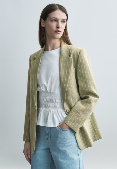 Femme aux longs cheveux bruns portant un blazer beige à fines rayures, un chemisier blanc froncé et un jean bleu clair, se tenant devant un fond uni gris clair.