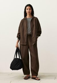 Žena v rozvolněném hnědém kabátě a kalhotách, pruhovaném tričku, držící černou tašku, stojící na světlé podlaze s neutrálním pozadím.