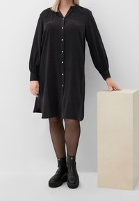 Femme portant une robe noire à boutons, des collants noirs transparent, et des bottes à plateforme noires, reposant une main sur un piédestal rectangulaire beige.