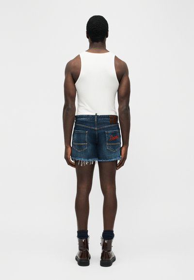 Άνδρας στέκεται και κοιτάζει μακριά, φοράει λευκό μπλουζάκι χωρίς μανίκια, ξεφτισμένα τζιν σορτσάκια με τη λέξη "Rebel" στην τσέπη, σκούρες κάλτσες και καφέ μποτάκια.