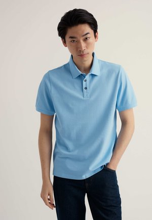 Mladić stoji s jednom rukom u džepu, nosi svijetloplavu polo majicu kratkih rukava i tamne traperice, na neutralnoj pozadini.