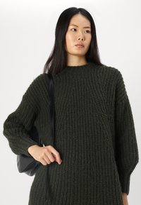 Maglione lavorato a maglia verde scuro con texture a coste, dotato di un collo alto e di una vestibilità comoda. Il modello tiene in mano una borsa nera con tracolla.