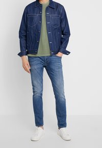 Homme portant une veste en denim bleu, un t-shirt vert olive, un jean bleu et des baskets blanches sur un fond blanc uni.