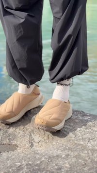 Beige slip-on sko med teksturert materiale og gummisåler, kombinert med hvite sokker og svarte bukser, plassert på en stein nær vann.