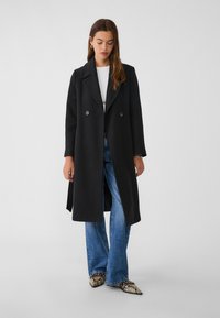 Svart ullkappa med dubbelknäppt design, inskärpt krage och sidofickor, i kombination med utsvängda blå denimjeans och mönstrade skor.
