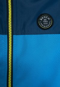 Niebieska kurtka z odważnym czarnym zamkiem błyskawicznym i okrągłą naszywką z napisem "BENCH ORIGINALS", w kolorowej kompozycji w granatowym i jasnoniebieskim kolorze.