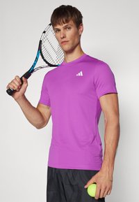 Camiseta deportiva morada de manga corta, con un tejido texturizado y un logo blanco. El modelo sostiene una raqueta de tenis con una mano y una pelota de tenis con la otra.