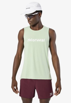 Hombre con camiseta sin mangas verde claro con texto "Nnormal", pantalones cortos burdeos, gorra blanca y gafas deportivas blancas, de pie contra un fondo liso.
