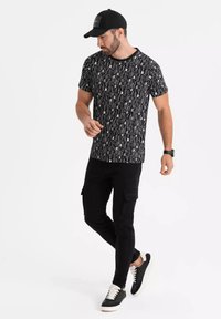 Camiseta gráfica negra con patrones verticales blancos, combinada con pantalones cargo negros y zapatillas negras con cordones blancos. Se incluye una gorra negra.