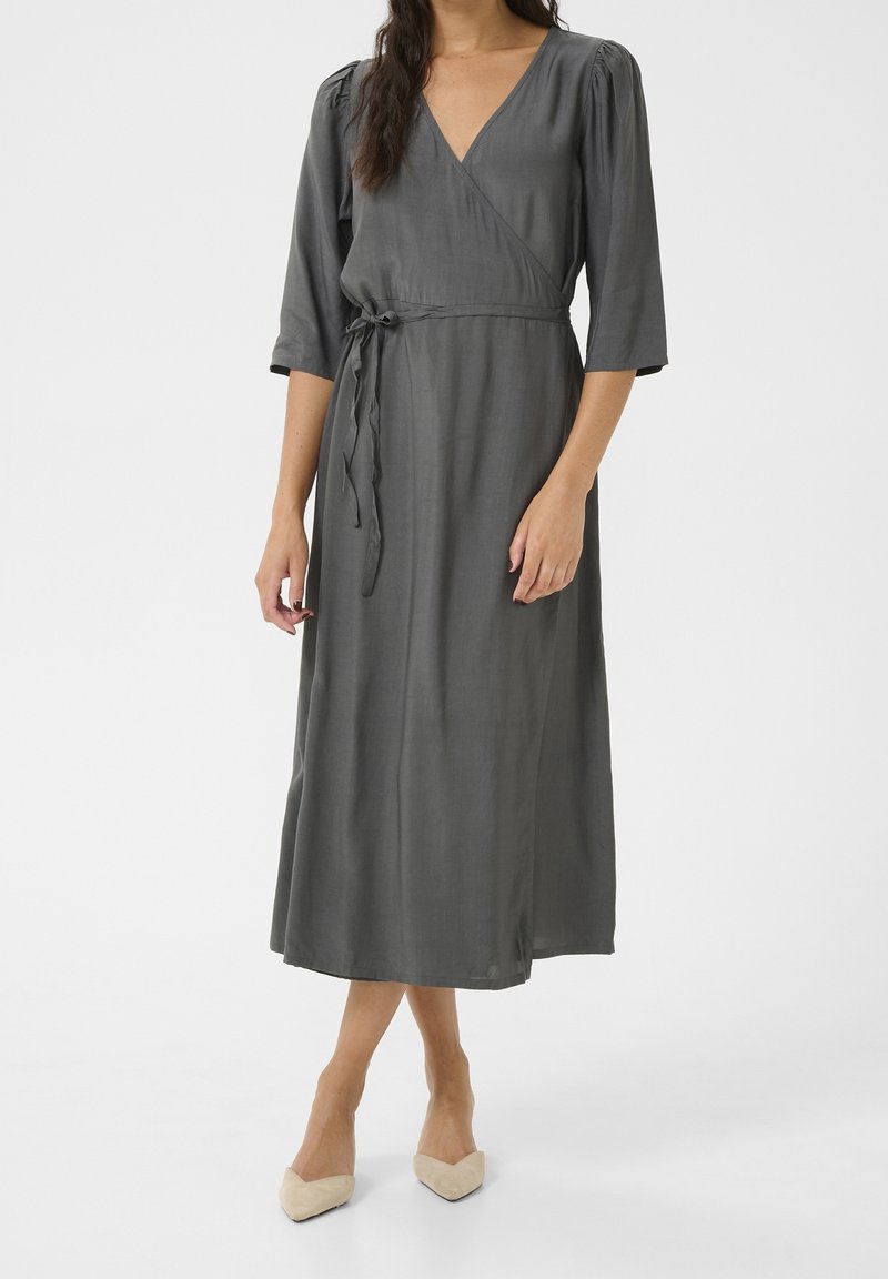 Femme portant une robe midi grise portefeuille à manches trois-quarts et des escarpins beiges à bouts pointus, debout devant un fond uni.