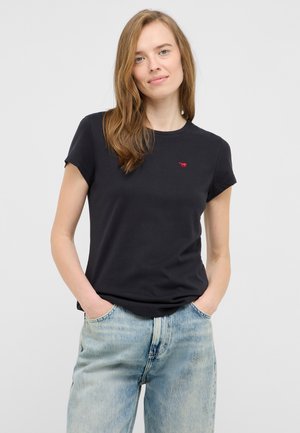Schwarzes Kurzarm-T-Shirt mit rundem Ausschnitt und kleinem roten Logo auf der Brust. Kombiniert mit hellblauen Jeans. Weiches Baumwollmaterial.