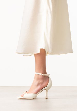 Vrouw draagt een offwhite enkelriem sandalen met hoge hak en strikdetail, gecombineerd met een vloeiende crèmekleurige midirok.