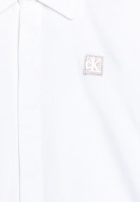 Camicia di cotone bianca con colletto classico e una piccola patch in argento con il logo "CK" sul lato sinistro superiore. Texture liscia e linee pulite.