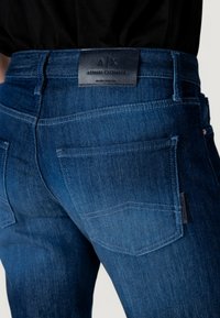 Jeans de mezclilla azul oscuro con un efecto desgastado, que cuentan con una etiqueta de cuero A/X en la cinturilla y dos bolsillos traseros con costuras distintas.