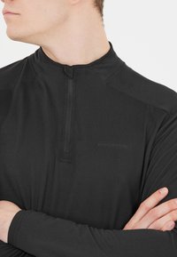 Sort langærmet bluse med en halv lynlås og en høj krave. Lavet af et glat, let materiale med subtil tekstur og branding.