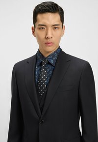 Completo nero con rever a punta, abbinato a una camicia a fiori blu e a una cravatta a motivo geometrico con forme a diamante. La trama appare liscia.