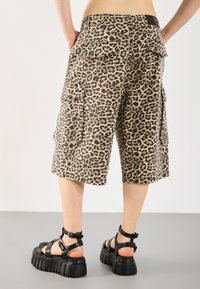 Beige krēslas ar leopardu rakstu, ar melniem akcentiem, ar lielām kabatām, brīvā piegriezumā, kopā ar masīviem melniem sandaliem.