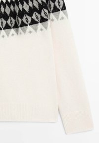 Manche de pull en tricot blanc et torse inférieur avec un motif de losanges noirs, gris et blancs près de l'épaule.