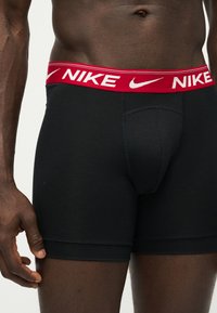 Ανδρικός κορμός με μαύρα μποξεράκια Nike με κόκκινη μέση που φέρει λευκό λογότυπο Nike και το σήμα swoosh, στέκεται σε απλό φόντο.