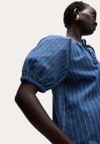 Blusa di denim blu con righe verticali bianche, maniche corte a sbuffo e dettaglio con fiocco al collo. Tessuto morbido, vestibilità comoda.