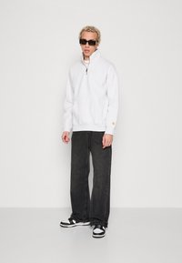 Hvid fleece sweatshirt med lynlås og høj krave, parret med løse sorte denimjeans og sorte og hvide sneakers. Mørke solbriller på.