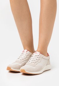 Sneakers beige realizzati in materiali mesh e scamosciati, con una suola bianca imbottita e accenti corallo sul tallone e sulla linguetta, lacci ben dettagliati.